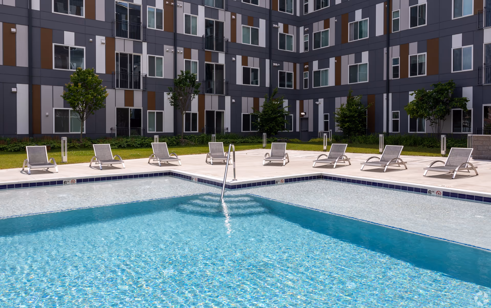 Outdoor swimming pool with clear blue water surrounded by a concrete deck with several lounge chairs. The pool area is enclosed by a modern multi-story building with a patterned facade in shades of gray and brown, and several small trees and greenery around the perimeter.
