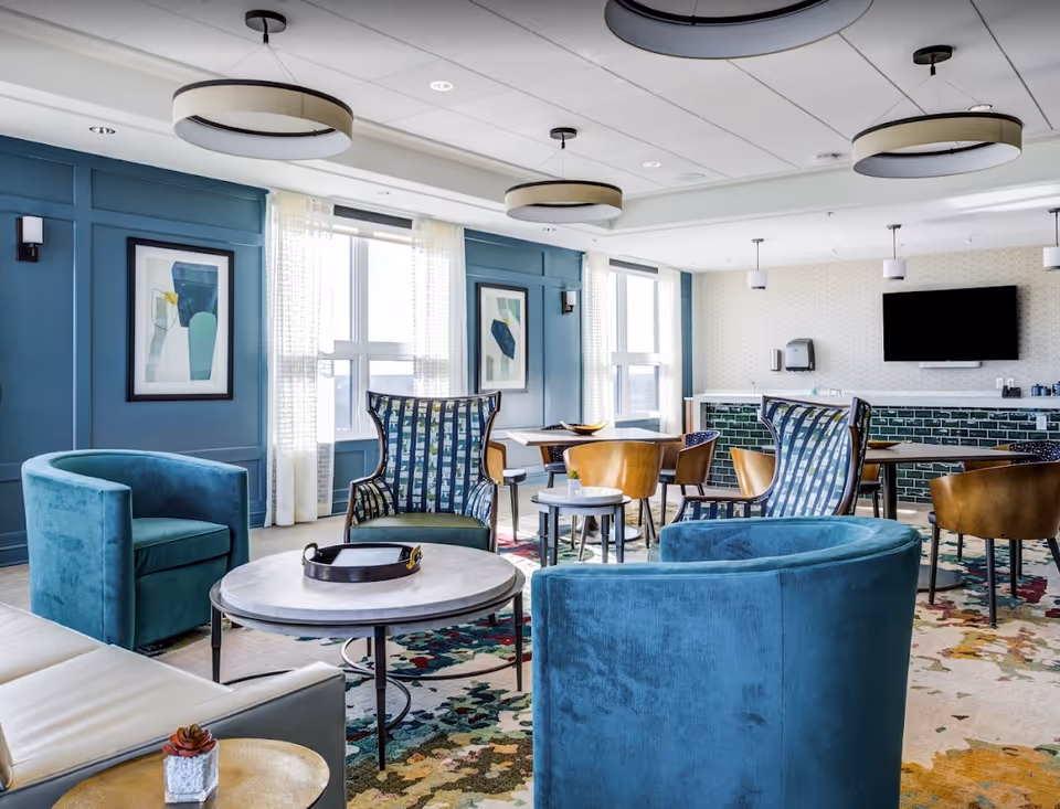 A bright and modern common area with blue and teal armchairs, round tables, and wooden chairs. The room features large windows with sheer curtains, blue paneled walls with framed abstract art, and a patterned carpet. There are circular ceiling lights and a wall-mounted TV above a counter with green tile backsplash.