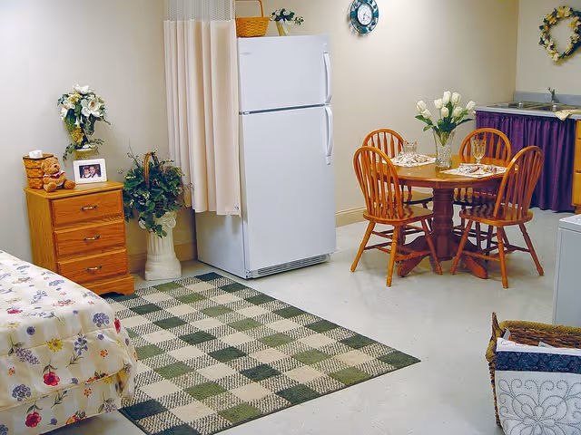 Studio-style interior with a bed and dresser to the left, a white refrigerator and potted plants in the center, and a round dining table with four wooden chairs to the right.