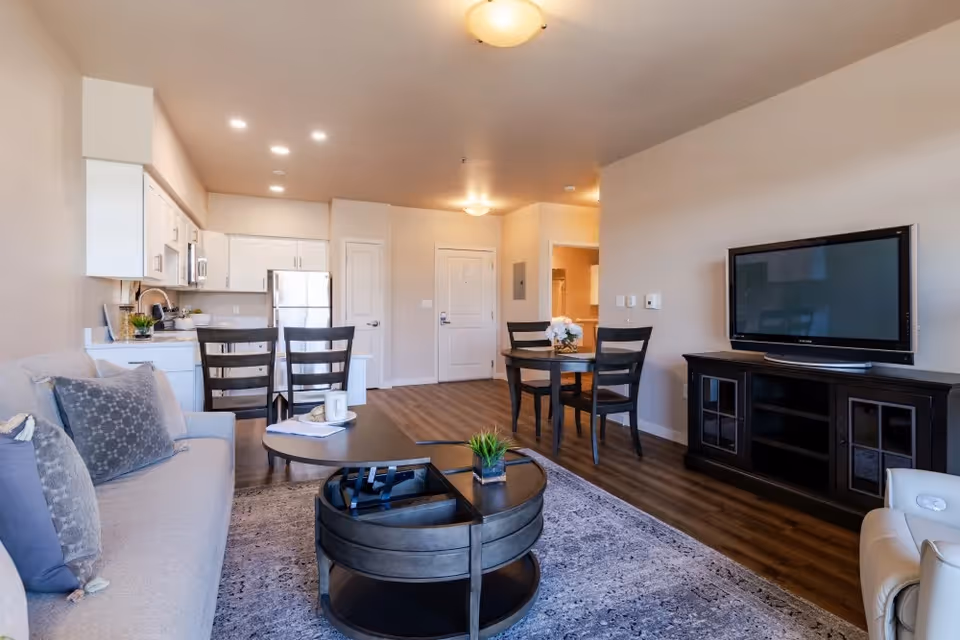 A bright and modern living area in a senior living facility featuring a beige sofa with decorative pillows, a round coffee table with a small plant and a cup, a flat-screen TV on a dark wooden stand, a dining table with four chairs, and a kitchen area with white cabinets and stainless steel appliances in the background.