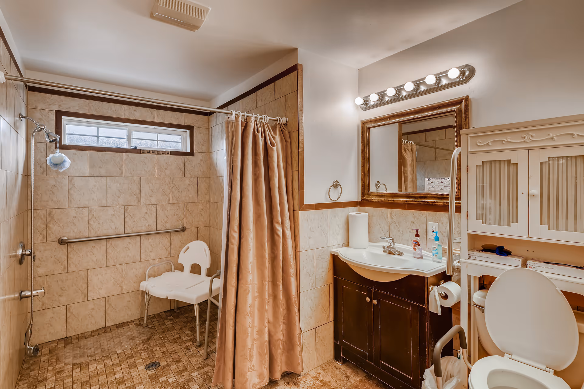 A bathroom with beige tiled walls and floor, featuring a walk-in shower area with a white shower chair and a grab bar. There is a beige shower curtain, a sink with a dark wooden cabinet below, a large framed mirror above the sink with multiple light bulbs, and a white cabinet next to the toilet. The toilet has armrests and a grab bar for accessibility.