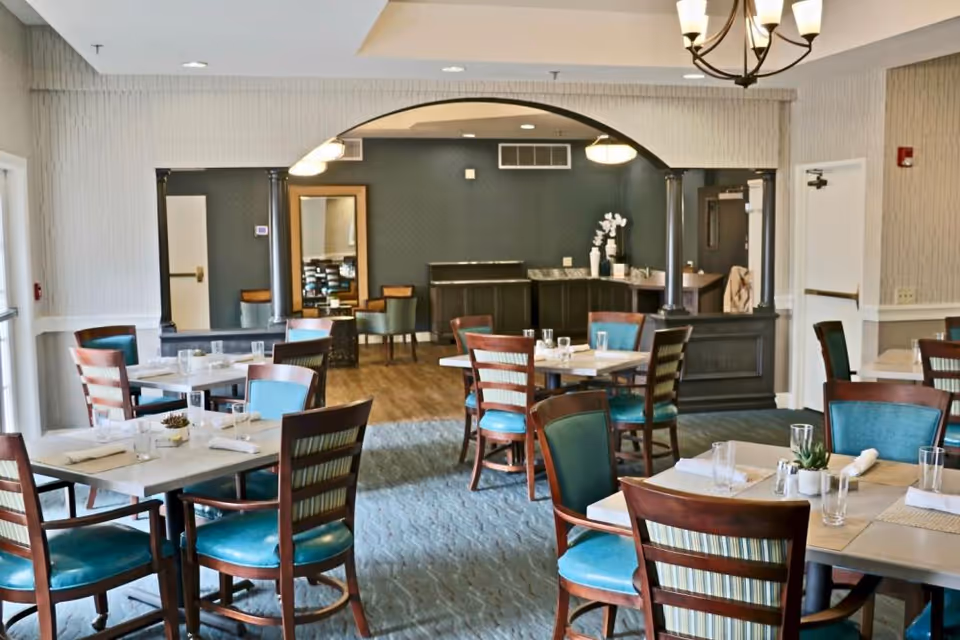 A dining room in a senior living facility with several tables and chairs arranged neatly. Each table is set with glasses, napkins, and small decorative plants. The room has a carpeted floor, light-colored walls, and a chandelier hanging from the ceiling. There is an archway leading to another room with additional seating and a counter with decorative items.