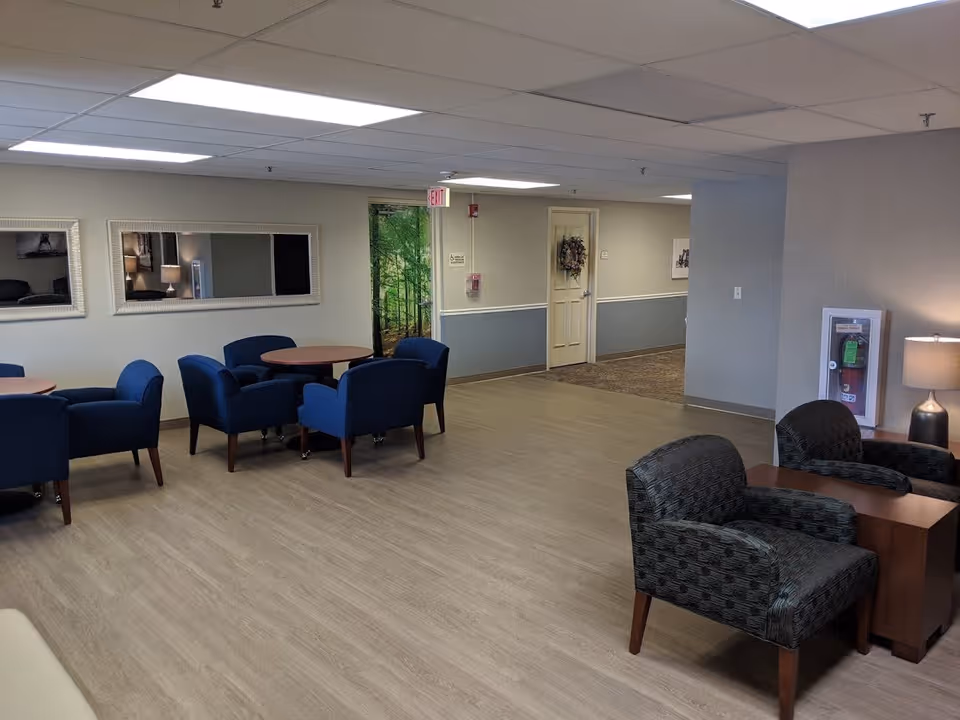 A spacious common area with multiple seating arrangements including blue upholstered chairs around round wooden tables and patterned armchairs around a small wooden table. The room has light wood flooring, neutral-colored walls with a chair rail, two large rectangular mirrors on one wall, a door decorated with a wreath, and a fire extinguisher cabinet with a lamp on a side table nearby. The ceiling has recessed lighting panels.