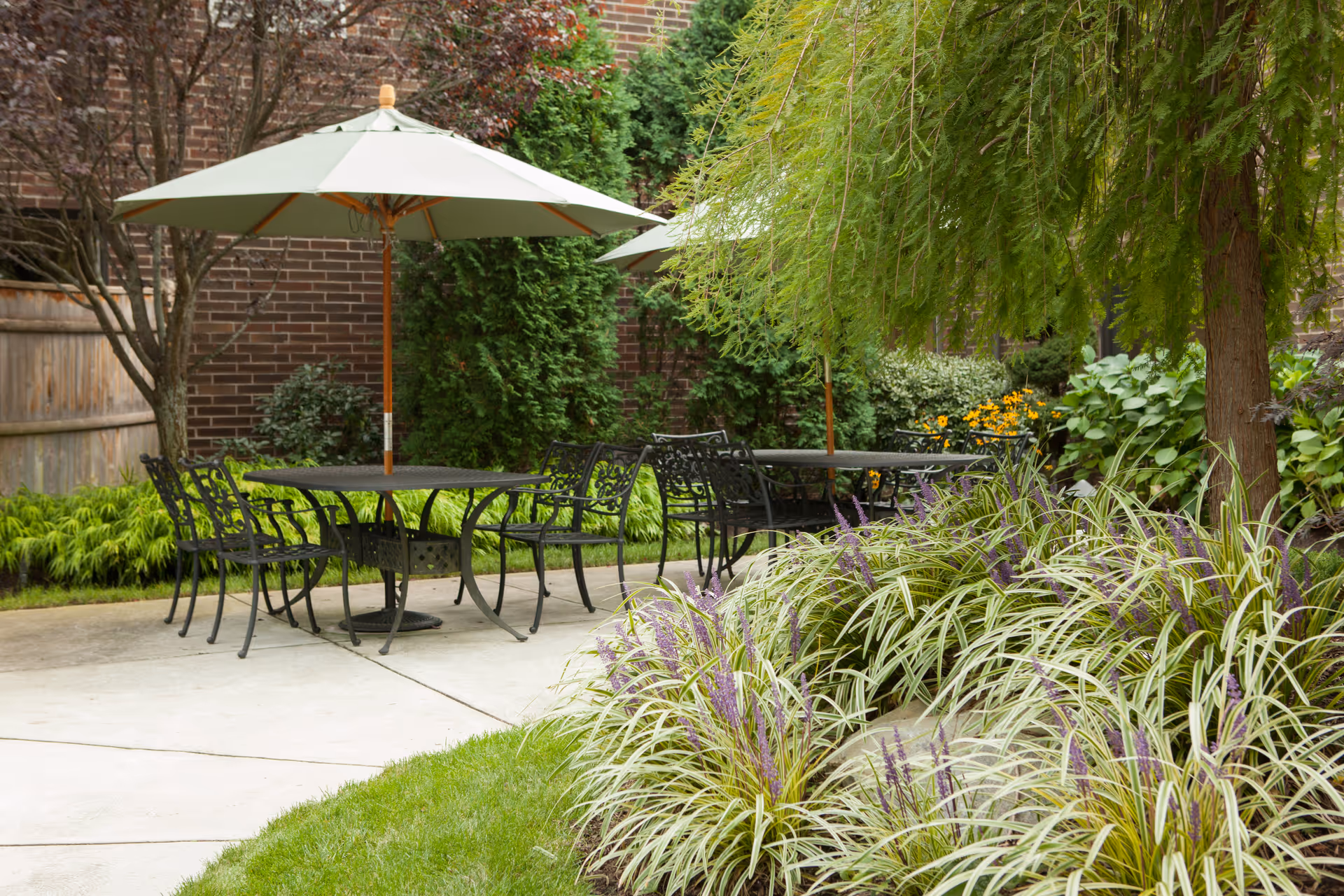 Outdoor patio area with black metal tables and chairs under large white umbrellas surrounded by green plants, trees, and a brick wall in the background.