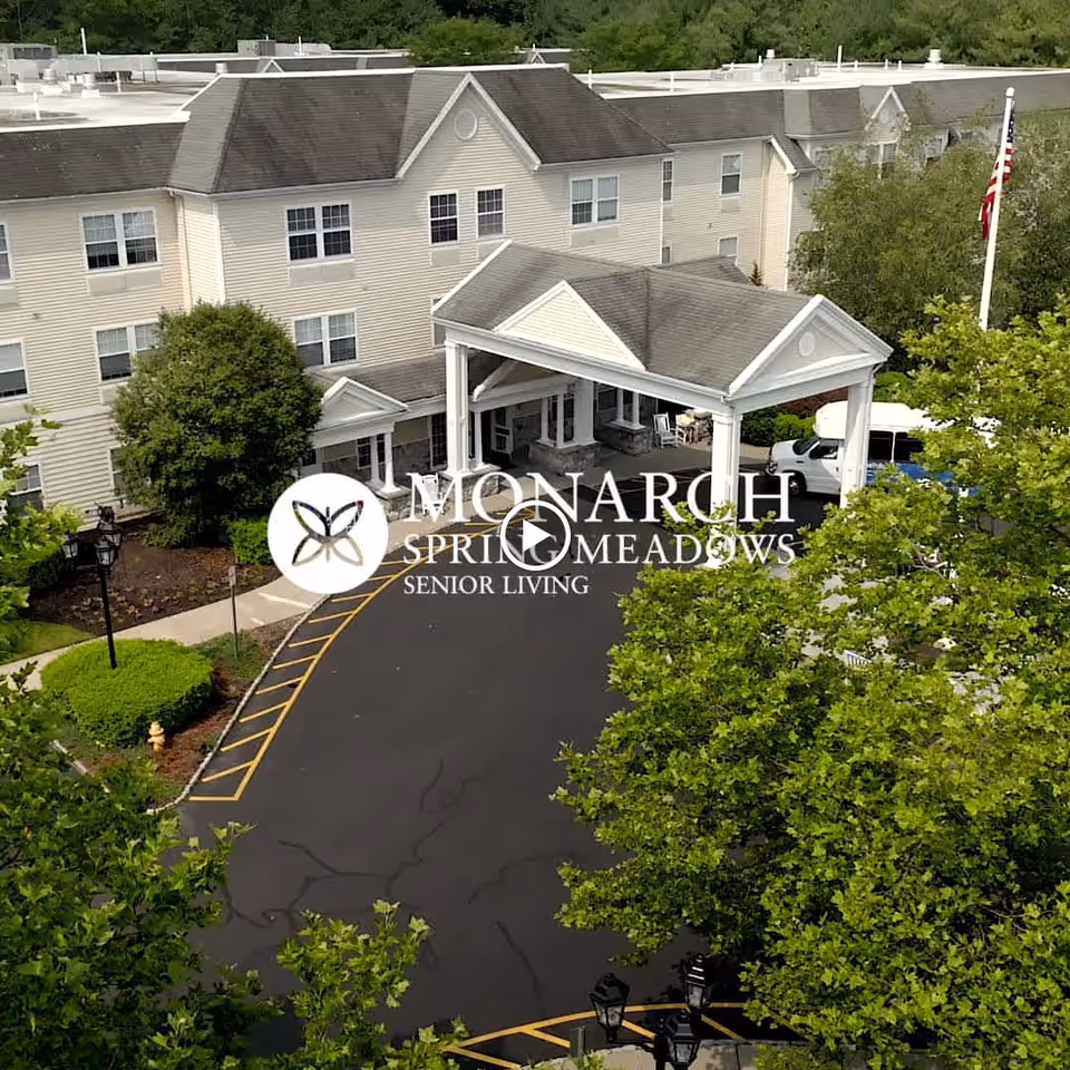 Exterior view of Monarch Spring Meadows senior living facility showing a large three-story building with beige siding, a covered entrance with white columns, a driveway with yellow parking lines, green trees, and an American flag on a flagpole.