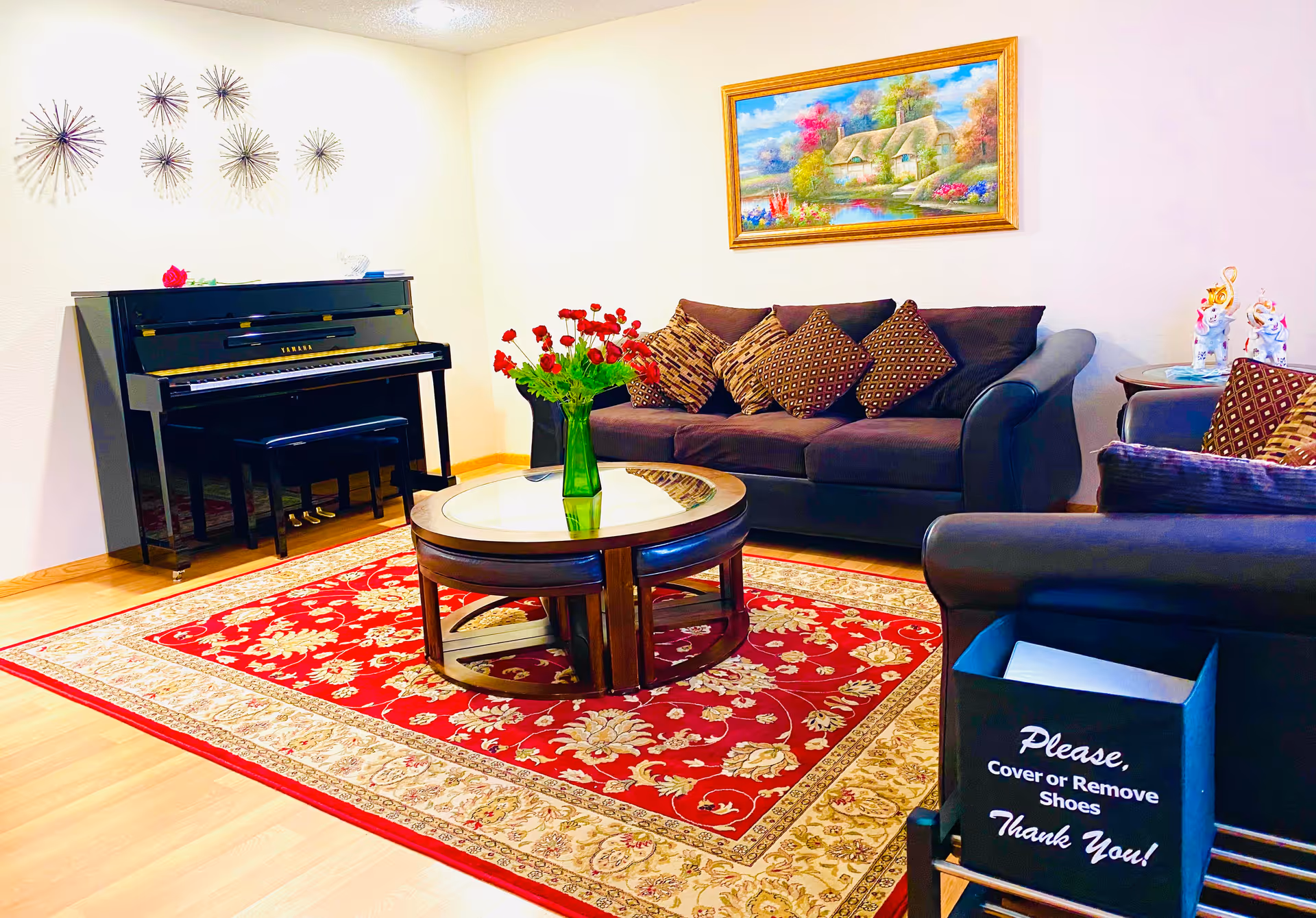 A cozy living room with a dark sofa and armchair adorned with patterned cushions, a round wooden coffee table with a glass top holding a green vase with red flowers, a red and beige floral area rug, a black upright piano against the wall, decorative wall art above the piano, and a framed painting of a cottage and garden on the wall.