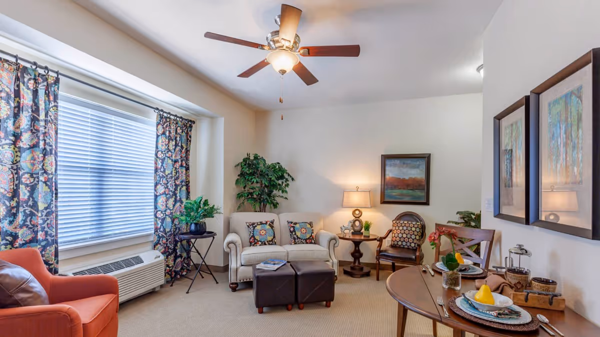 A cozy living room with a beige loveseat adorned with patterned pillows, an orange armchair, and a wooden chair with a patterned cushion. There is a round wooden dining table set with plates, utensils, and a yellow pear centerpiece. The room features a ceiling fan with a light, a large window with floral curtains, a potted plant, a side table with a lamp, and framed artwork on the walls.