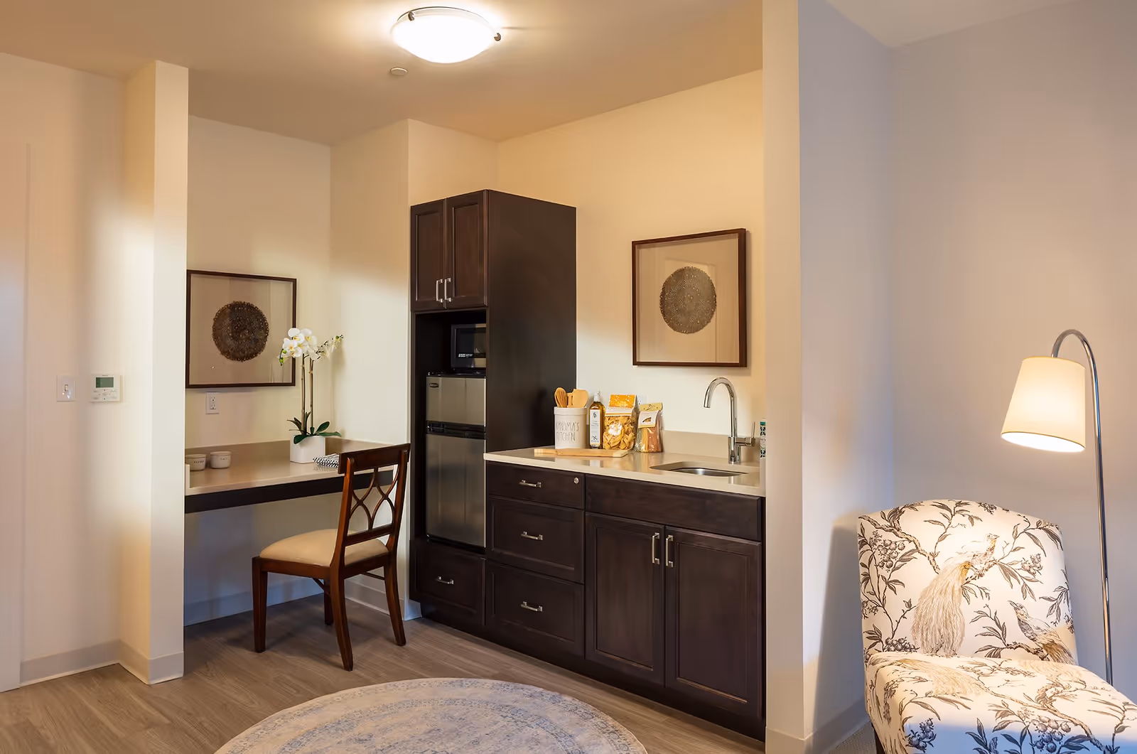 A cozy interior corner of a senior living residence featuring a small kitchenette with dark wood cabinets, a mini fridge, a microwave, and a sink. Next to the kitchenette is a built-in desk with a wooden chair and a potted orchid. On the right side, there is a floral-patterned armchair illuminated by a floor lamp. The walls are light-colored and decorated with framed artwork.