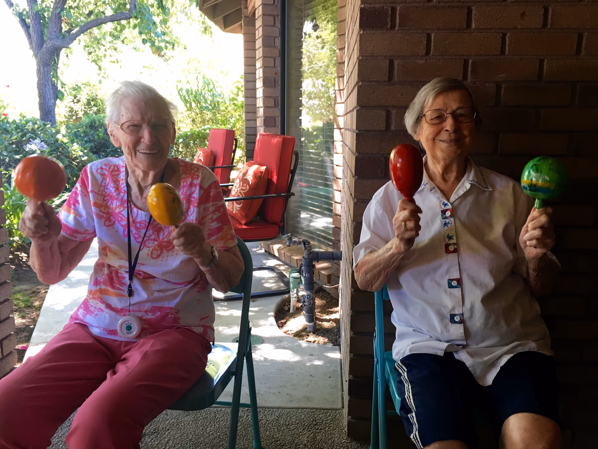 Two elderly women sitting on chairs outside on a shaded patio, each holding colorful maracas and smiling. Behind them are red cushioned chairs and green bushes with trees in the background.