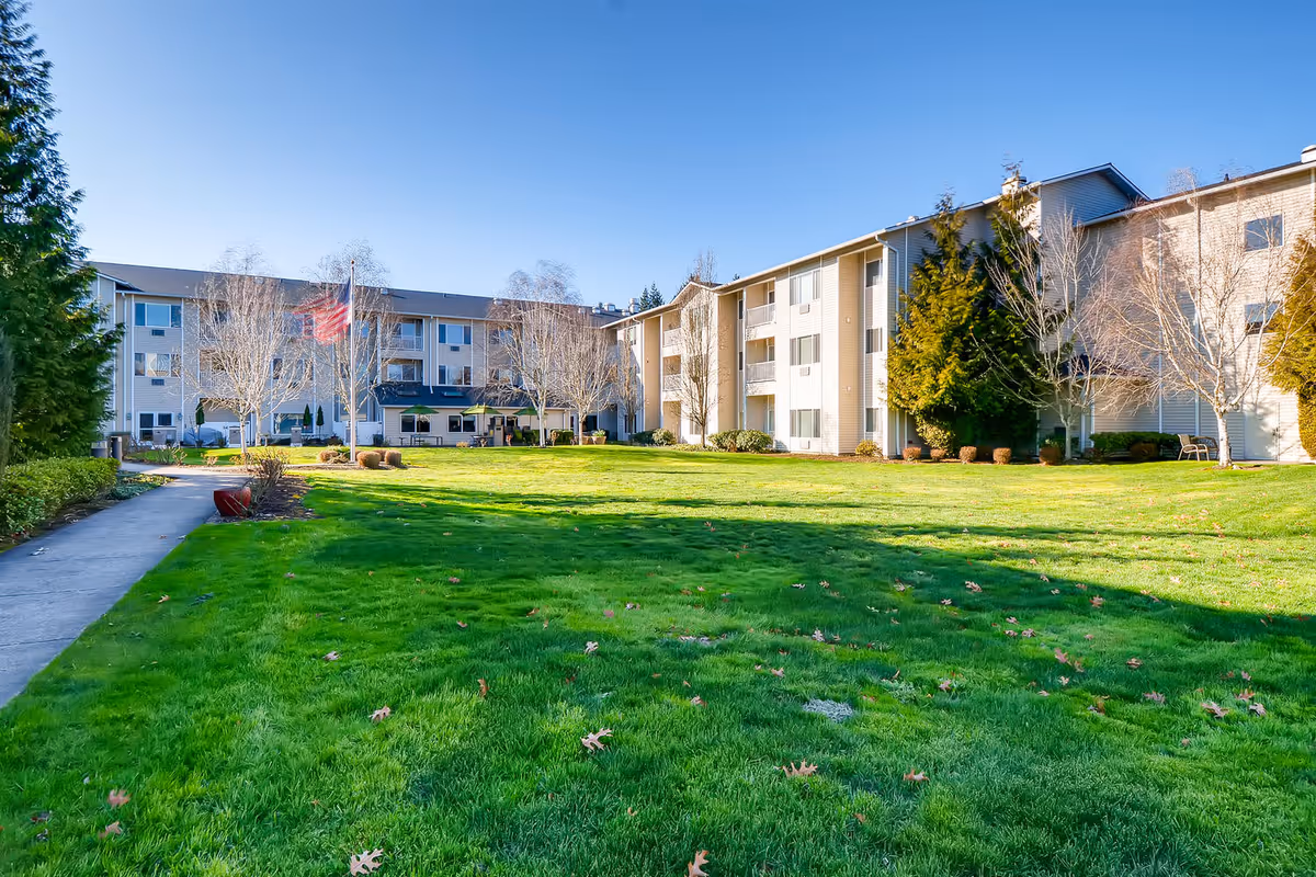A large green lawn with scattered fallen leaves in front of a three-story senior living facility building under a clear blue sky. There is a concrete walkway on the left side, some leafless trees, and an American flag on a flagpole near the center of the image.