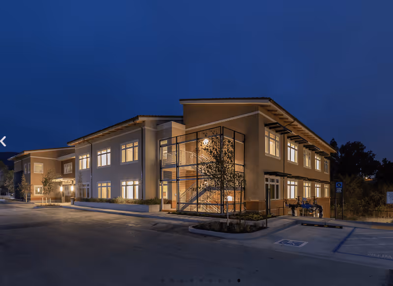 Exterior view of a two-story building at dusk with lights on inside. The building has large windows, a visible outdoor staircase enclosed in glass, and a parking area with handicap spaces in front. Trees and landscaping surround the building.