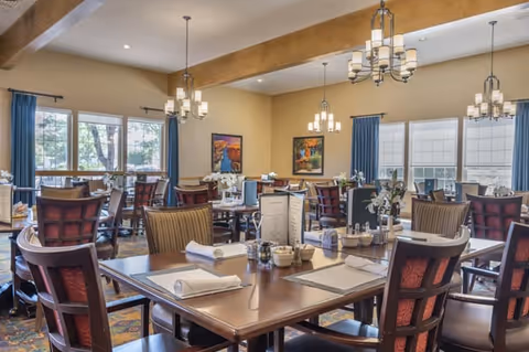A spacious dining room with multiple wooden tables and chairs arranged neatly. Each table is set with white napkins, menus, and small flower arrangements. Large windows with blue curtains allow natural light to fill the room. Elegant chandeliers hang from the ceiling, and framed artwork decorates the beige walls.