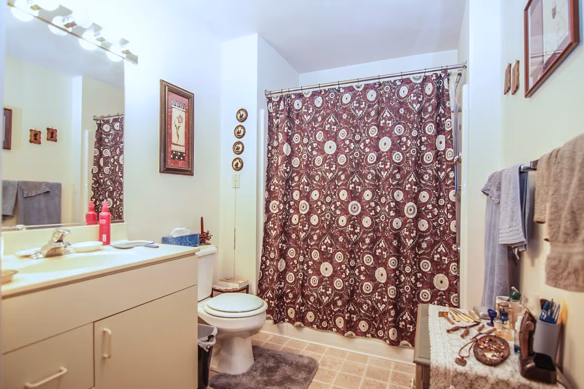 A bathroom with a white vanity and sink on the left, a toilet in the center, and a bathtub with a patterned maroon and white shower curtain. The walls are light-colored with framed artwork and decorative plates. Towels hang on a rack on the right wall, and various toiletries and personal items are placed on a small table covered with a lace cloth.