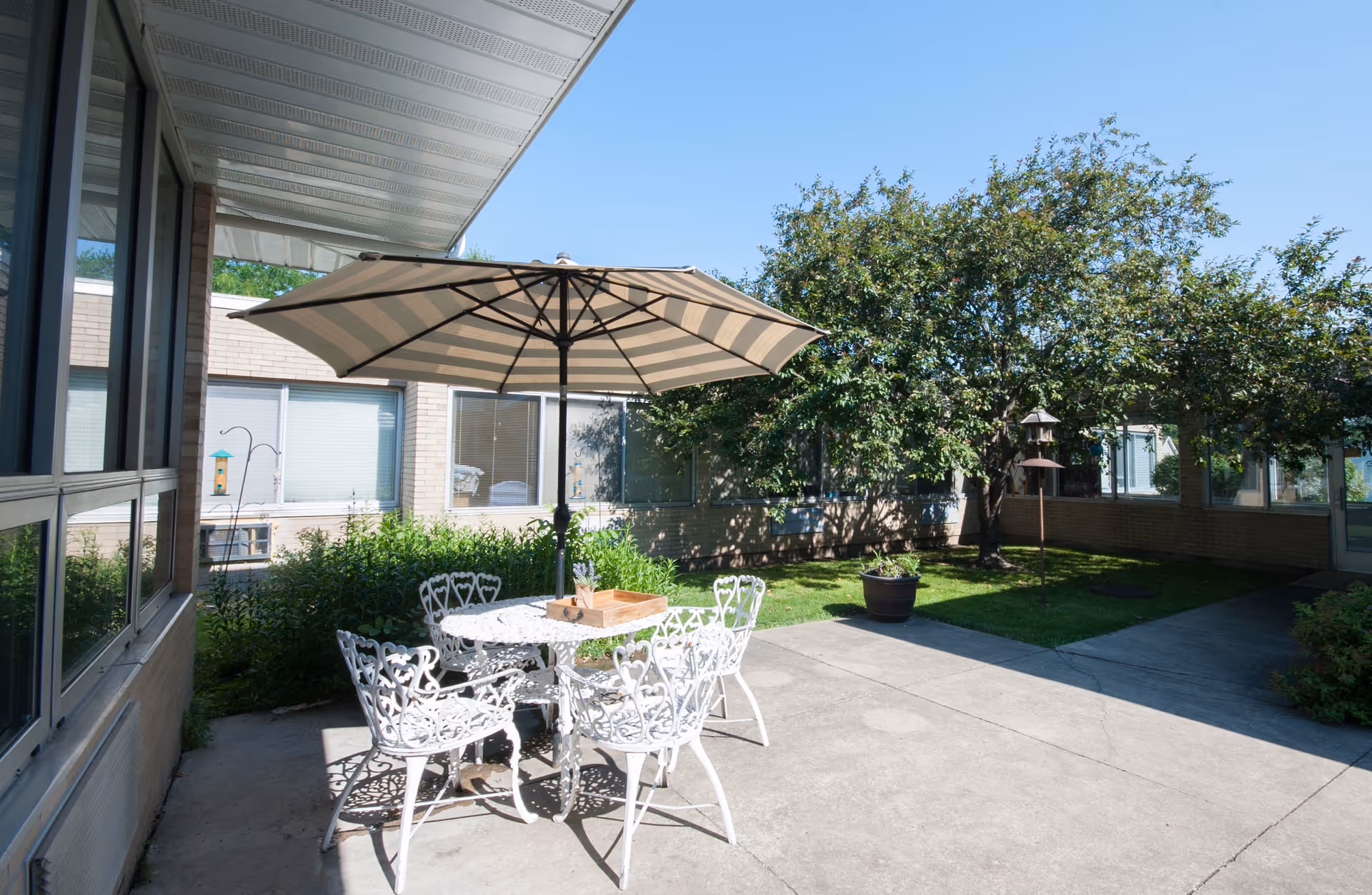 Outdoor patio area at a senior living facility with a white metal table and four matching chairs under a large striped umbrella. The patio is surrounded by a building with windows and greenery, including a tree and potted plants, under a clear blue sky.