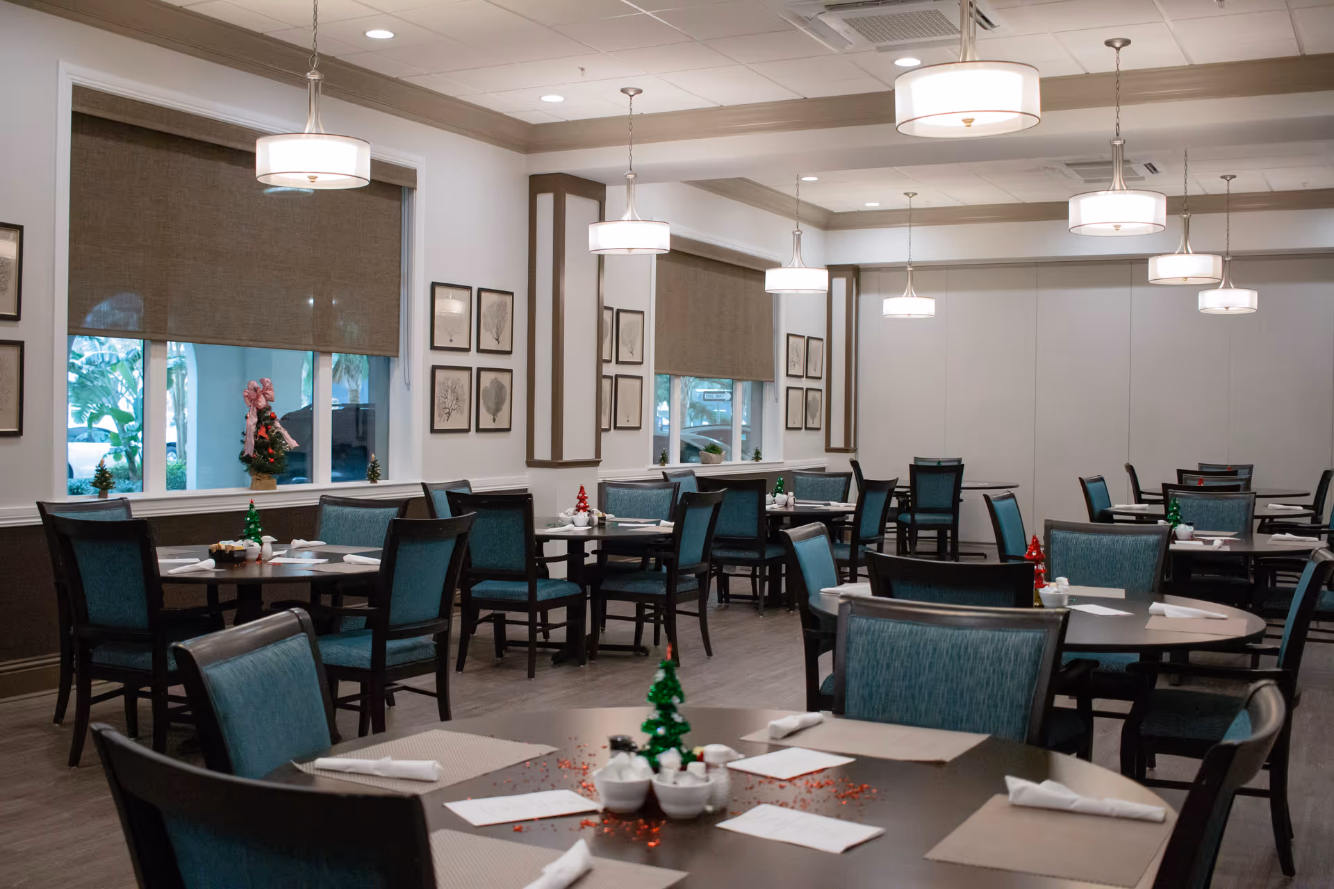 A spacious dining room with multiple tables and chairs arranged neatly. Each table has placemats, napkins, menus, and small holiday-themed centerpieces. The room features large windows with brown roller shades, framed botanical artwork on the walls, and modern pendant lights hanging from the ceiling. The floor is wood, and the overall decor is clean and inviting.