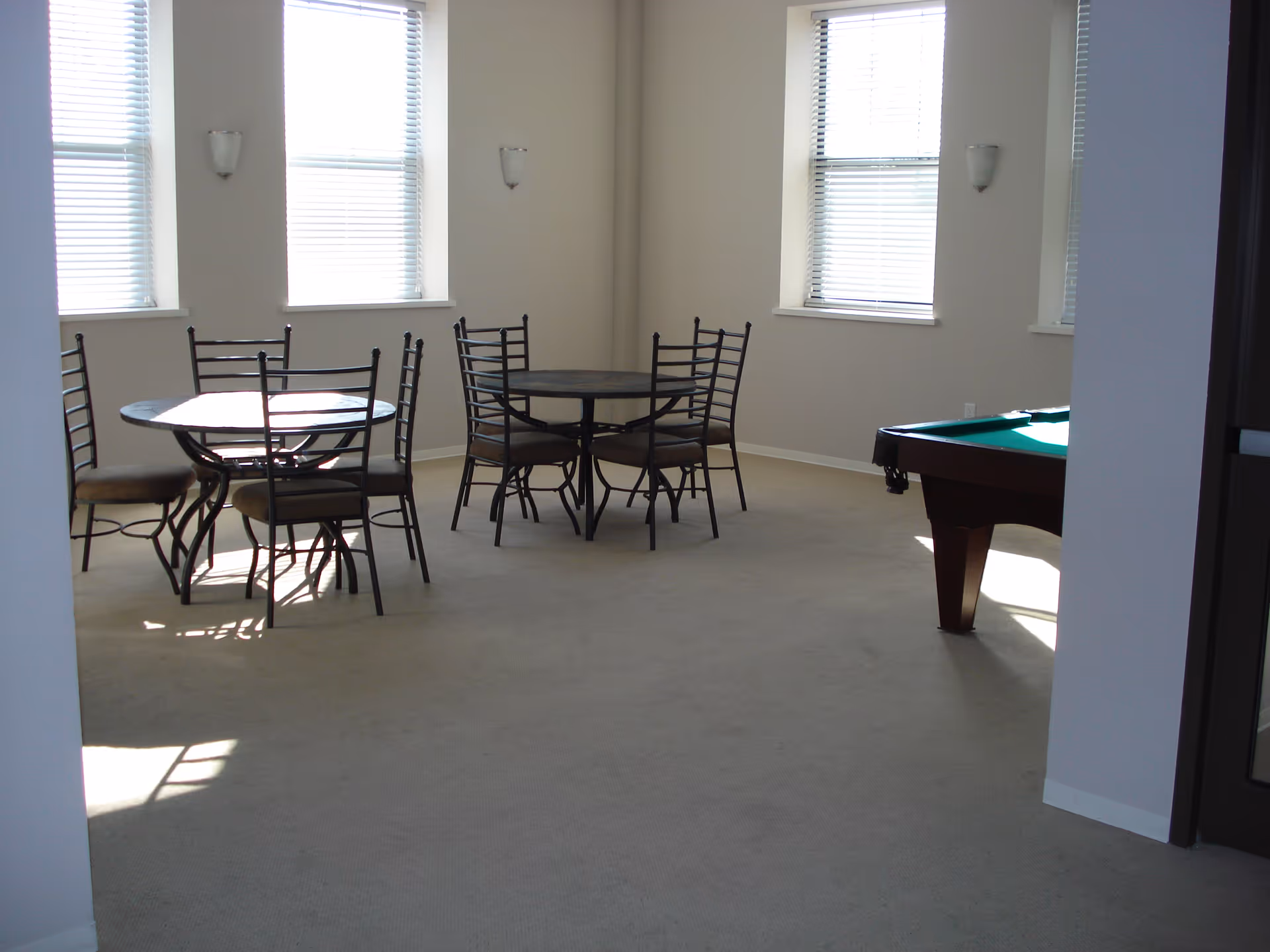 A bright room with beige carpet and several windows with blinds. There are two round tables each surrounded by four metal chairs with cushioned seats. A pool table is partially visible on the right side of the room.