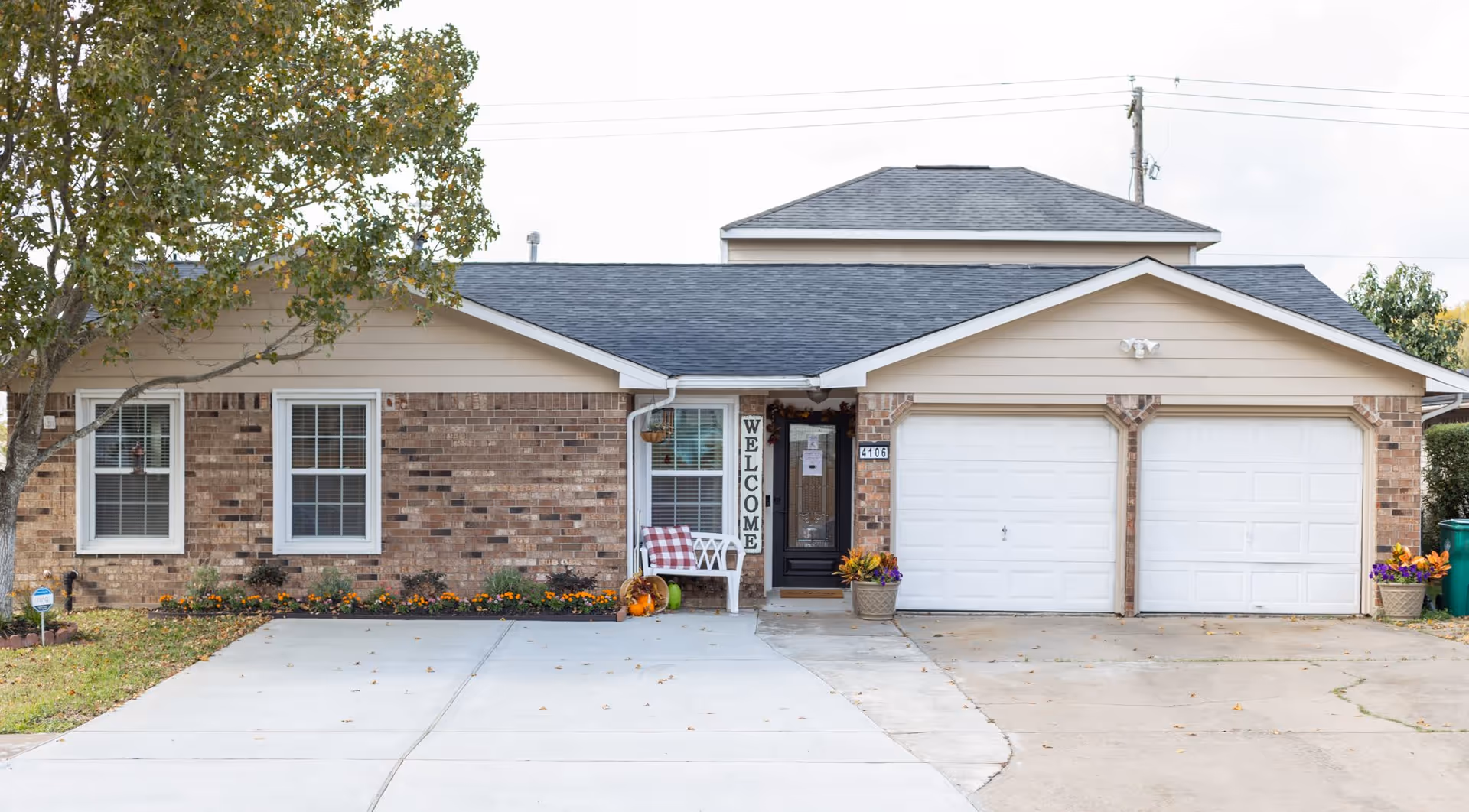 Front exterior view of a single-story brick house with a black front door, two white garage doors, a white bench with a red and white checkered cushion, and a vertical 'WELCOME' sign next to the door. There are flower pots with colorful flowers on either side of the garage and near the entrance, a tree on the left side, and a concrete driveway in front.