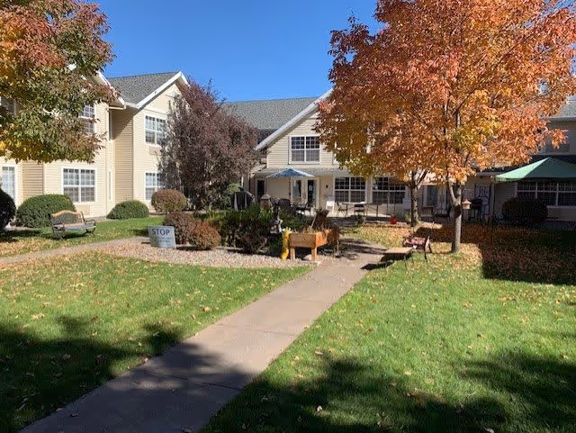 Outdoor courtyard area of McKay Creek Assisted Living with a paved walkway leading to the building entrance. The scene includes green grass, autumn-colored trees, benches, and a small garden area under a clear blue sky.