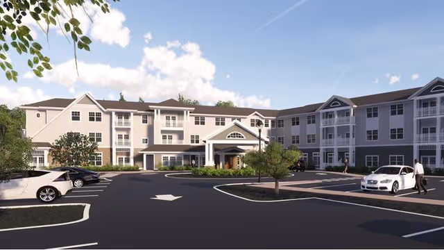 Exterior view of a three-story retirement community building with multiple windows and balconies. The building has a covered entrance with columns and a circular driveway in front. Several cars are parked in the parking lot, and a few people are walking near the entrance. The sky is partly cloudy with blue skies.
