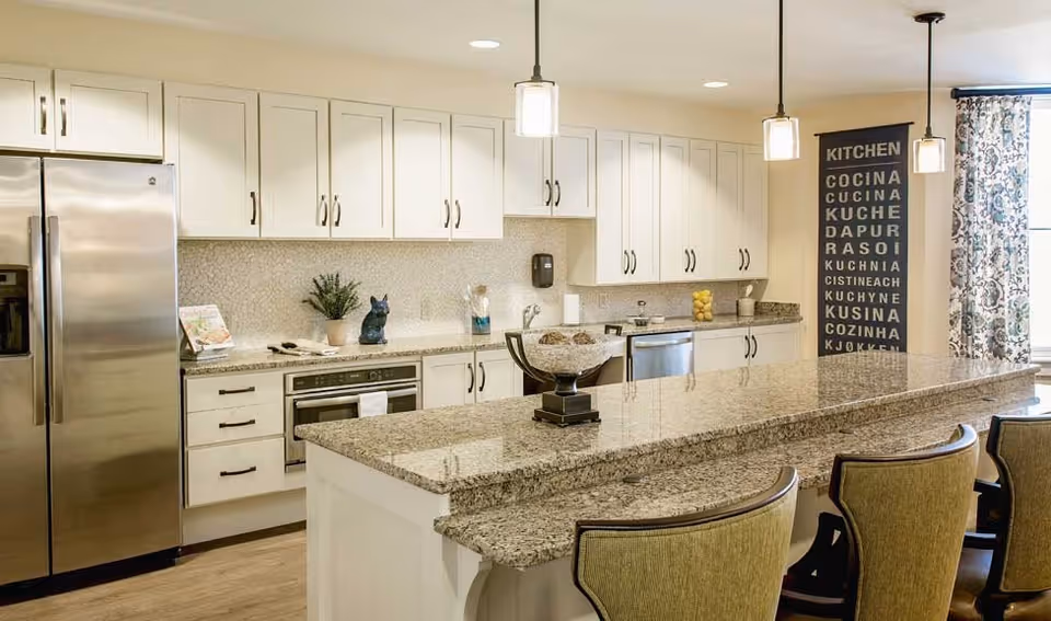 A modern kitchen with white cabinets, granite countertops, and stainless steel appliances including a refrigerator and oven. There is a large kitchen island with three upholstered chairs. Pendant lights hang above the island. A decorative sign with the word 'KITCHEN' in multiple languages is displayed on the wall near a window with patterned curtains.