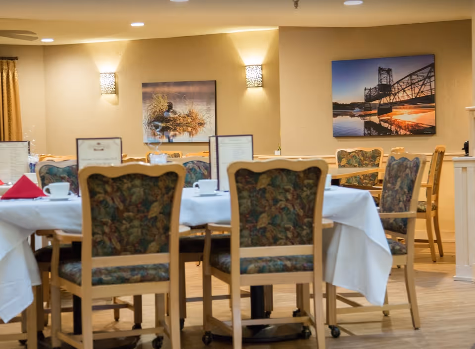 Dining room with tables covered in white tablecloths, set with cups, saucers, and menus. Wooden chairs with floral upholstery surround the tables. Two framed pictures hang on the beige walls, one depicting a duck on a nest and the other a bridge at sunset. Soft wall lights illuminate the room.