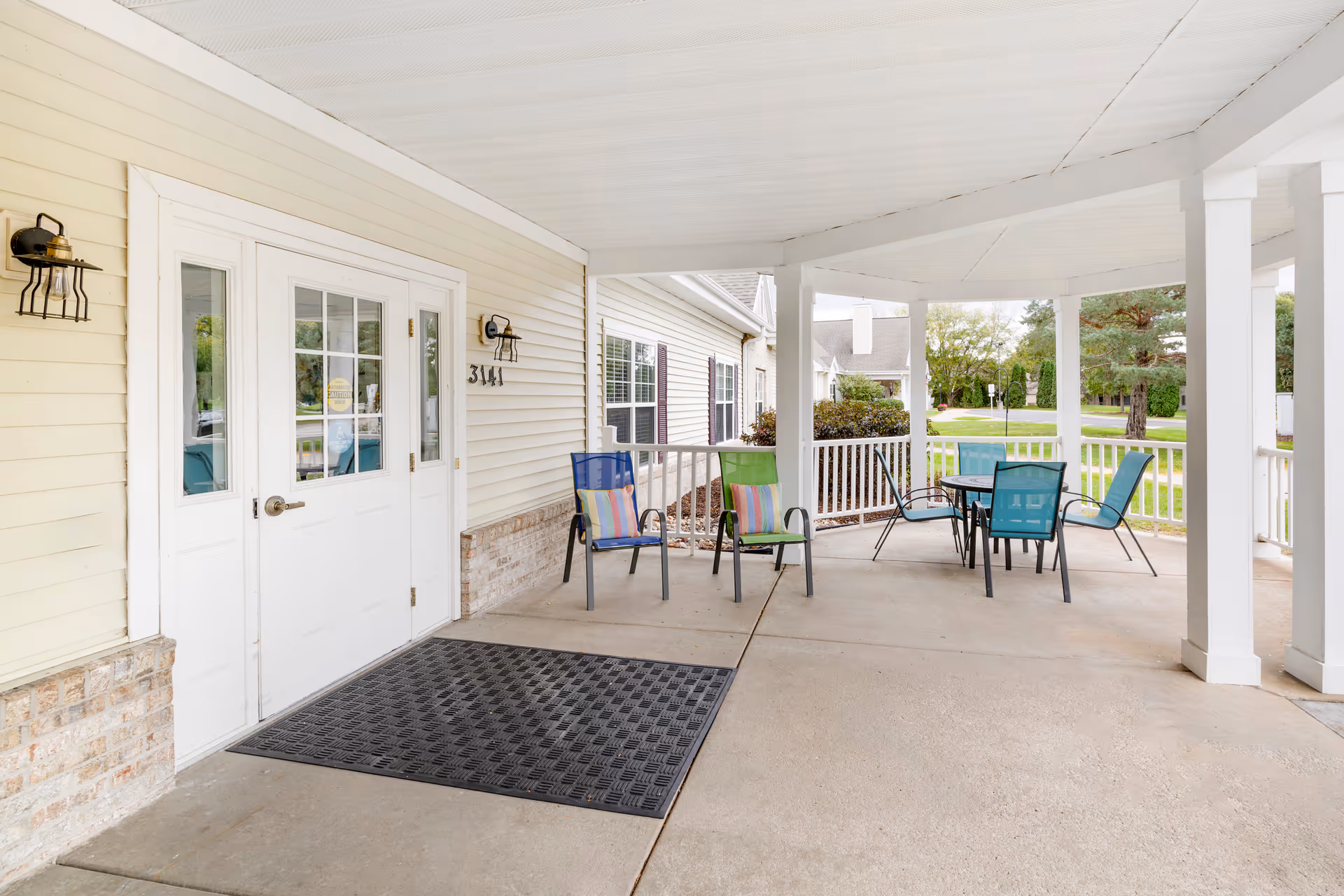 Covered outdoor patio area at Brookdale LaCrosse featuring white pillars and ceiling, a black rubber mat in front of white double doors, and several colorful chairs and a table arranged for seating. The background shows greenery and a street view.