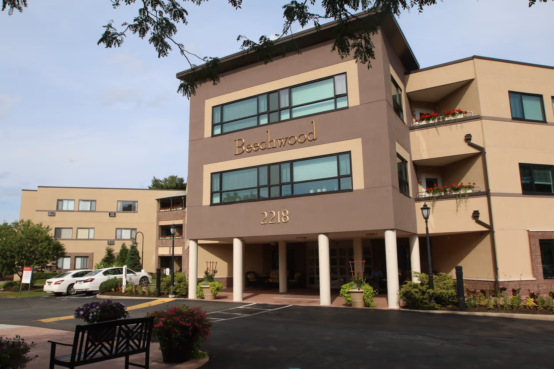 Exterior view of Beechwood at Eddy Memorial senior living facility showing a multi-story building with beige and brown walls, large windows, balconies with flower pots, a covered entrance supported by white columns, a parking area with cars, and landscaped greenery including potted plants and flowers.