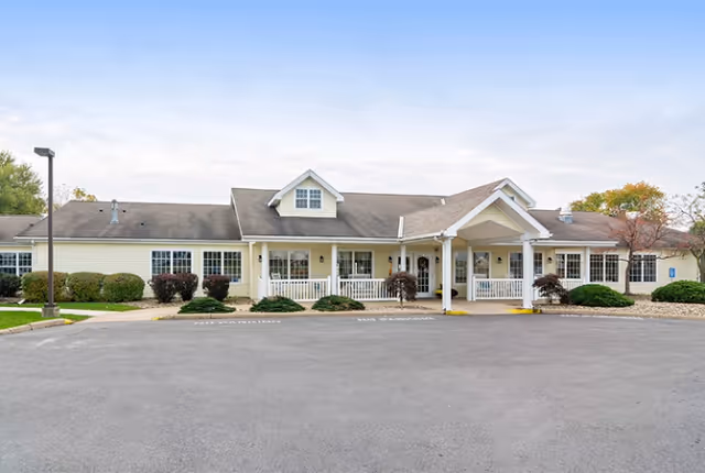 Front exterior view of a single-story senior living facility building with a covered entrance, white railings, and several windows. There are shrubs and small trees planted around the building and a large paved parking area in front.