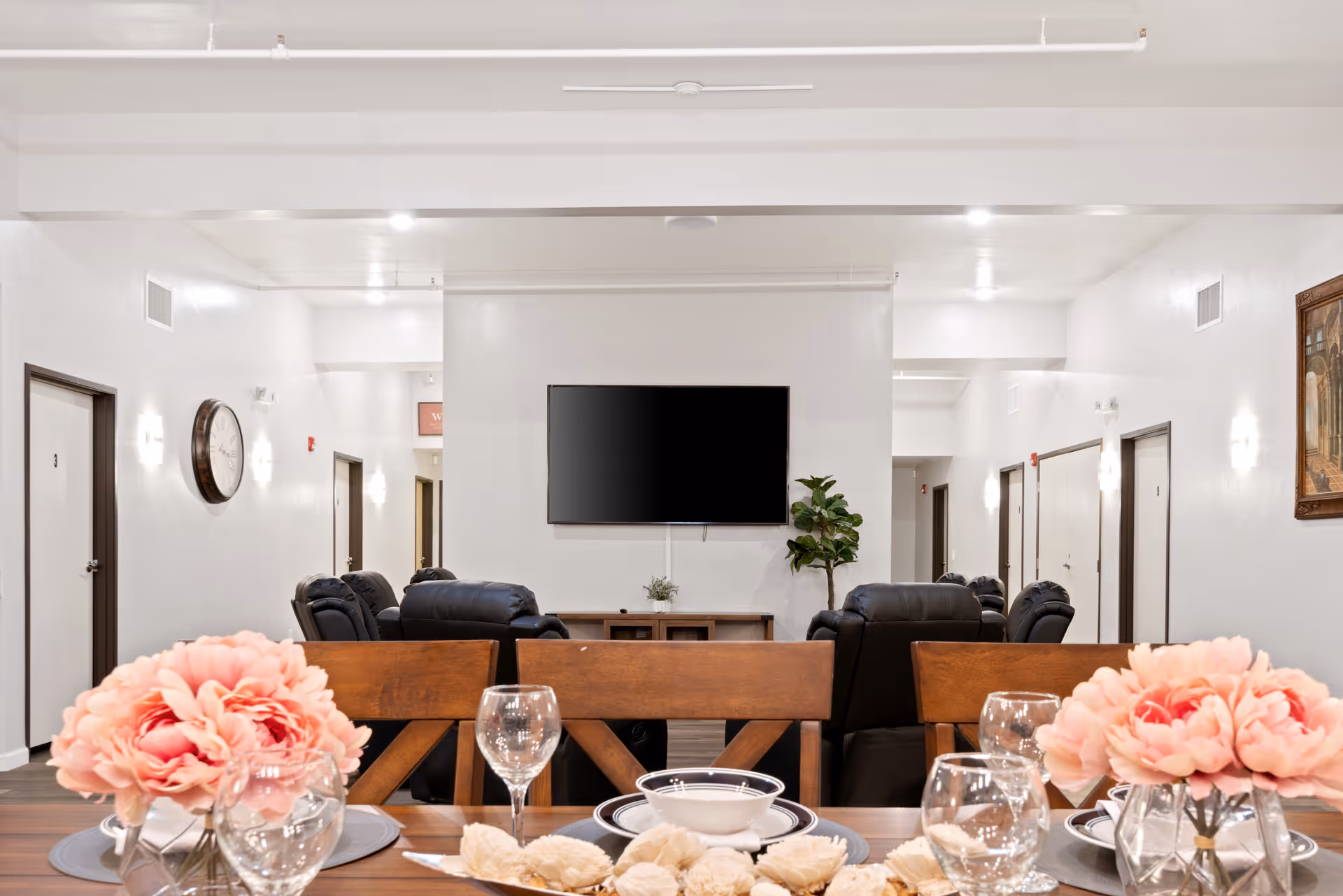 A senior living facility common area with a dining table in the foreground set with plates, bowls, glasses, and pink flower centerpieces. In the background, there are black leather recliners arranged facing a wall-mounted flat-screen TV, with a small plant and a cabinet beneath it. The room has white walls, several doors, a large wall clock, and a framed painting.