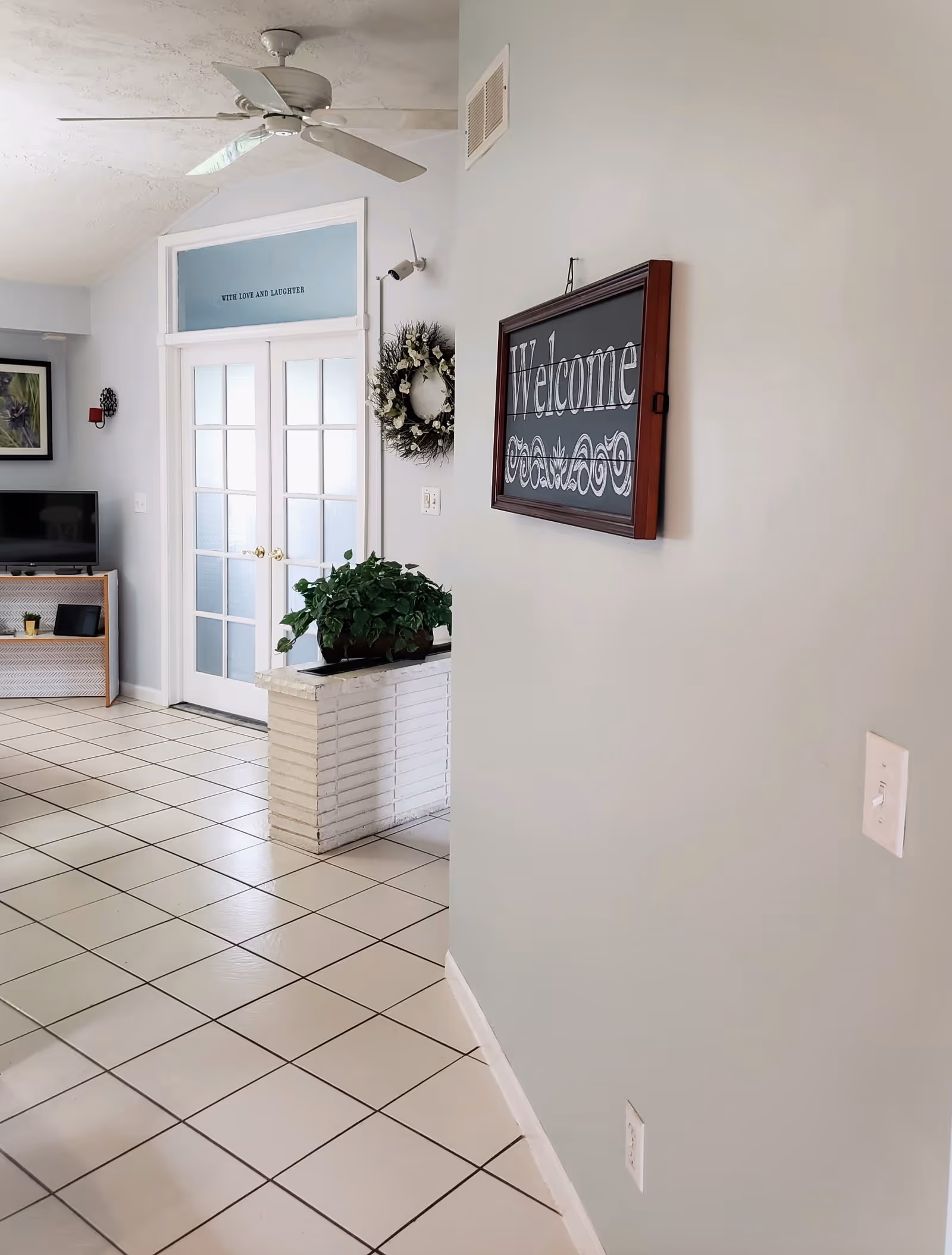 Interior view of a living area with light-colored tiled floor, a ceiling fan, a wall-mounted 'Welcome' sign, a plant on a white brick planter, and double glass doors with a sign above them that reads 'WITH LOVE AND LAUGHTER'. A TV on a wooden stand and a framed picture are visible in the background.