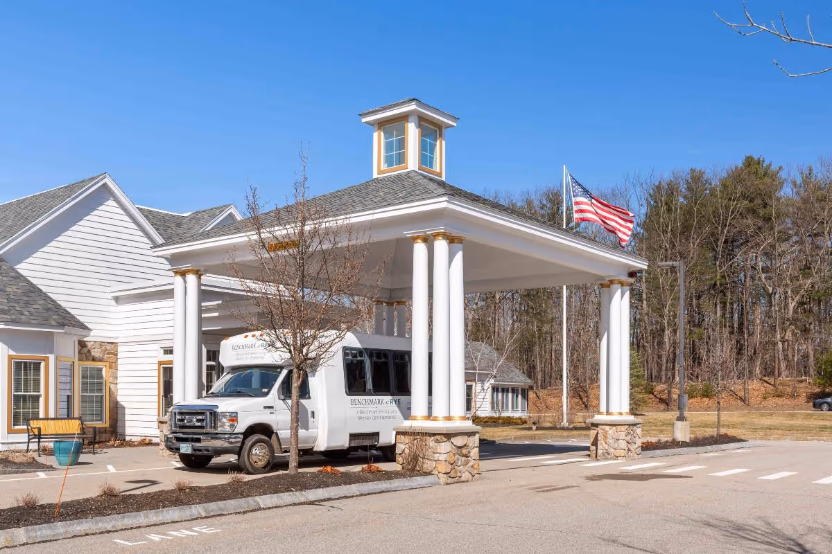 Entrance of a senior living facility named Benchmark at Rye with a covered drop-off area supported by white columns with gold accents. A white shuttle bus is parked under the canopy. An American flag is flying on a flagpole nearby, and there are trees and a clear blue sky in the background.