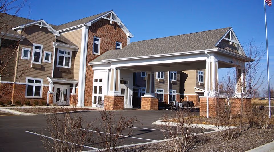 Exterior view of a senior living facility building with a covered entrance supported by white columns and brick bases. The building has beige siding with white trim and multiple windows. There is a parking area and some leafless shrubs in the foreground under a clear blue sky.