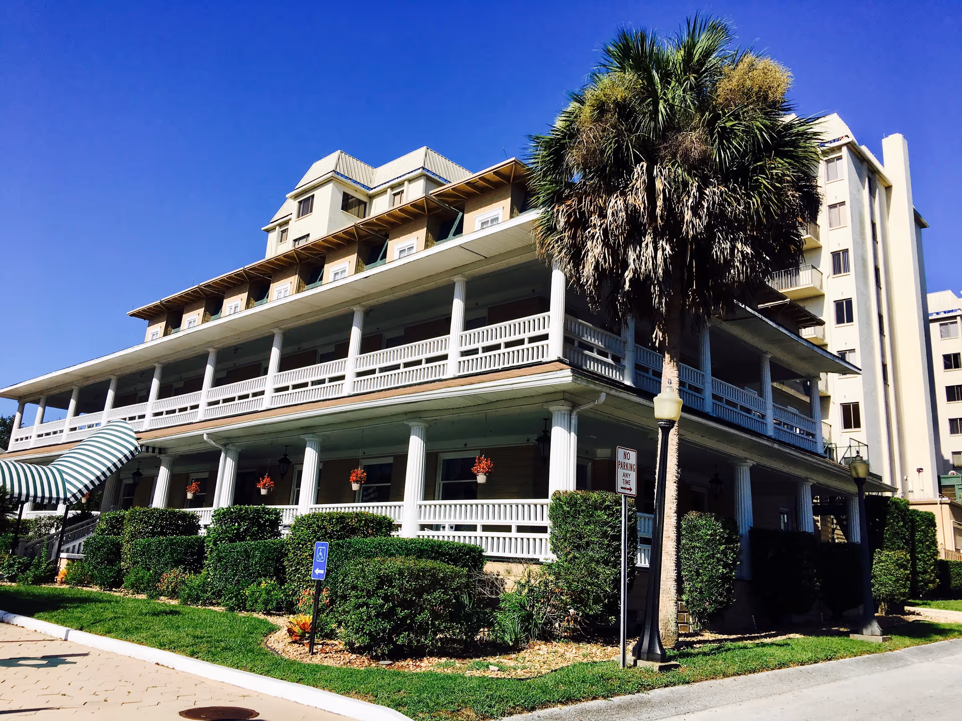 Exterior view of a multi-story building with white columns and railings, surrounded by green bushes and a palm tree, under a clear blue sky.