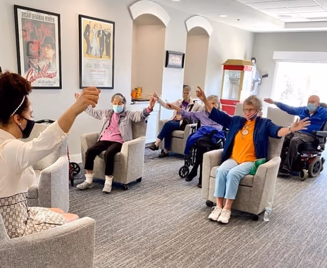 A group of elderly individuals seated in armchairs and wheelchairs in a common room, participating in a seated exercise session led by an instructor. Everyone is wearing face masks. The room has framed movie posters on the wall and a popcorn machine in the background.