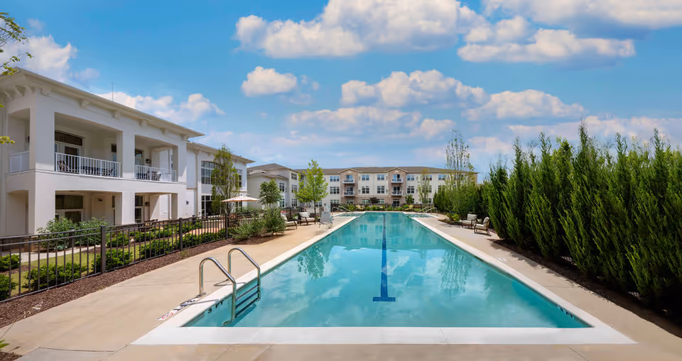Outdoor swimming pool with clear blue water surrounded by a concrete deck, lounge chairs, and greenery. On the left side, there is a white multi-story building with balconies, and in the background, there is another multi-story building under a partly cloudy blue sky.