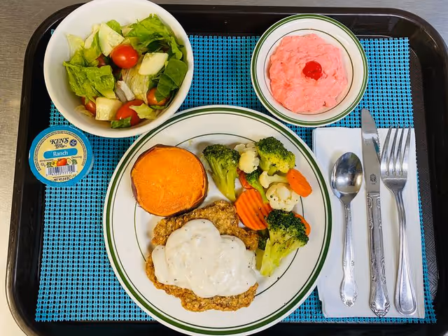 Tray with a plate of country-fried steak topped with gravy, steamed vegetables and a sweet potato, plus a side salad, pink dessert, utensils, and a cup of ranch dressing.