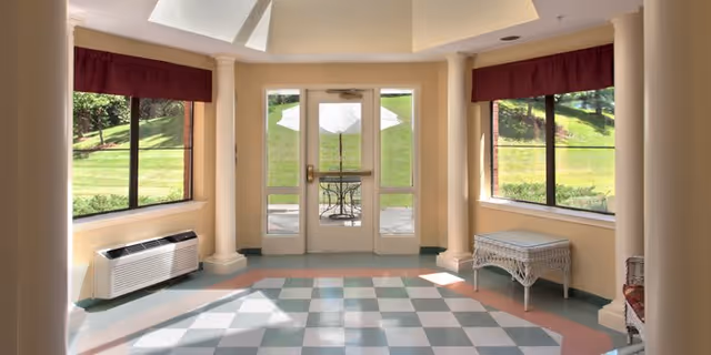 A bright interior room with large windows on either side and a glass door leading outside to a patio area with a table and umbrella. The room has checkered tile flooring, beige walls, white columns, and maroon valances above the windows. Outside, green grass and trees are visible.