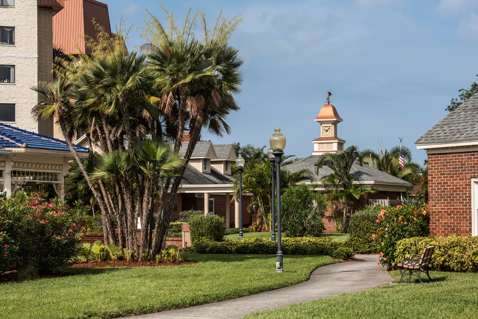 Landscaped courtyard with palm trees, a curved walkway, lamp posts and brick buildings with a clock tower.