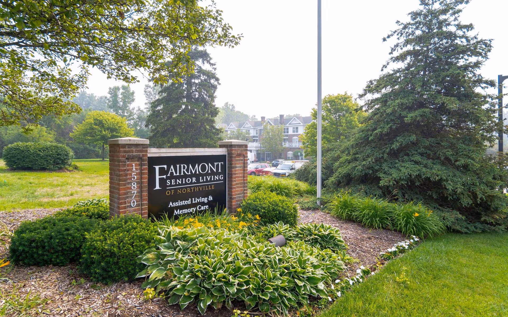Brick entry sign reading "Fairmont Senior Living of Northville" surrounded by landscaped shrubs and flowers with the facility building and parked cars visible behind it.