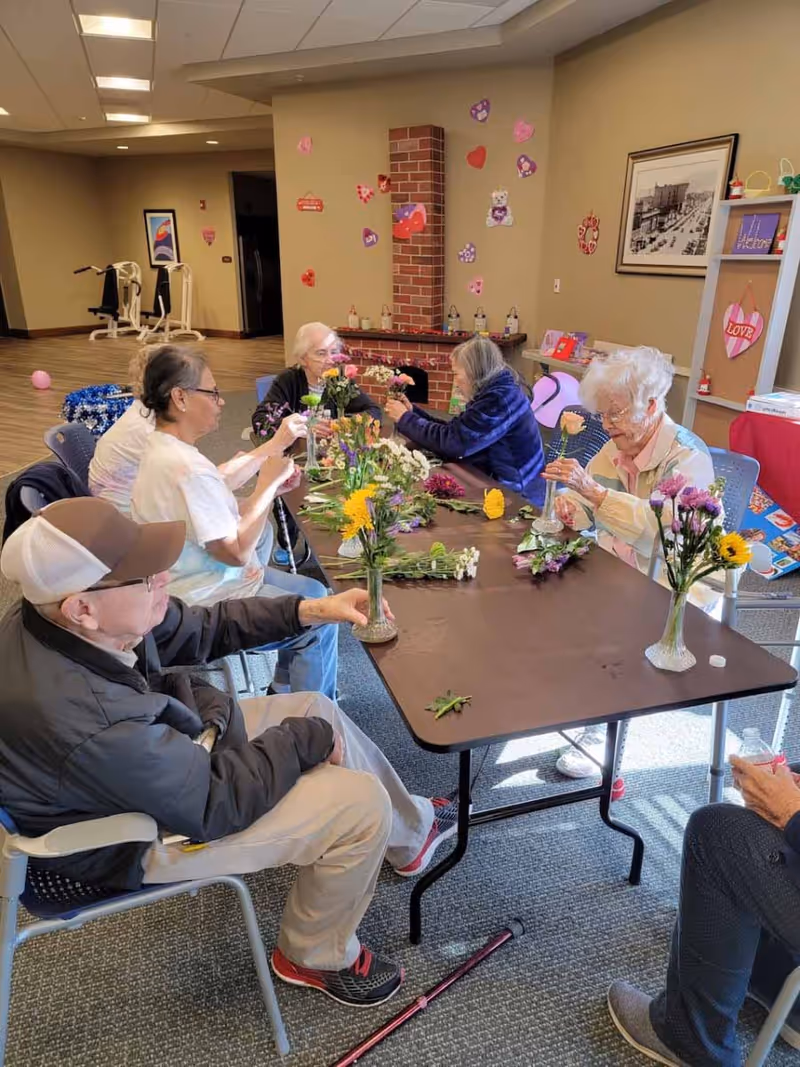Several residents seated around a table arranging flowers in a decorated common room.