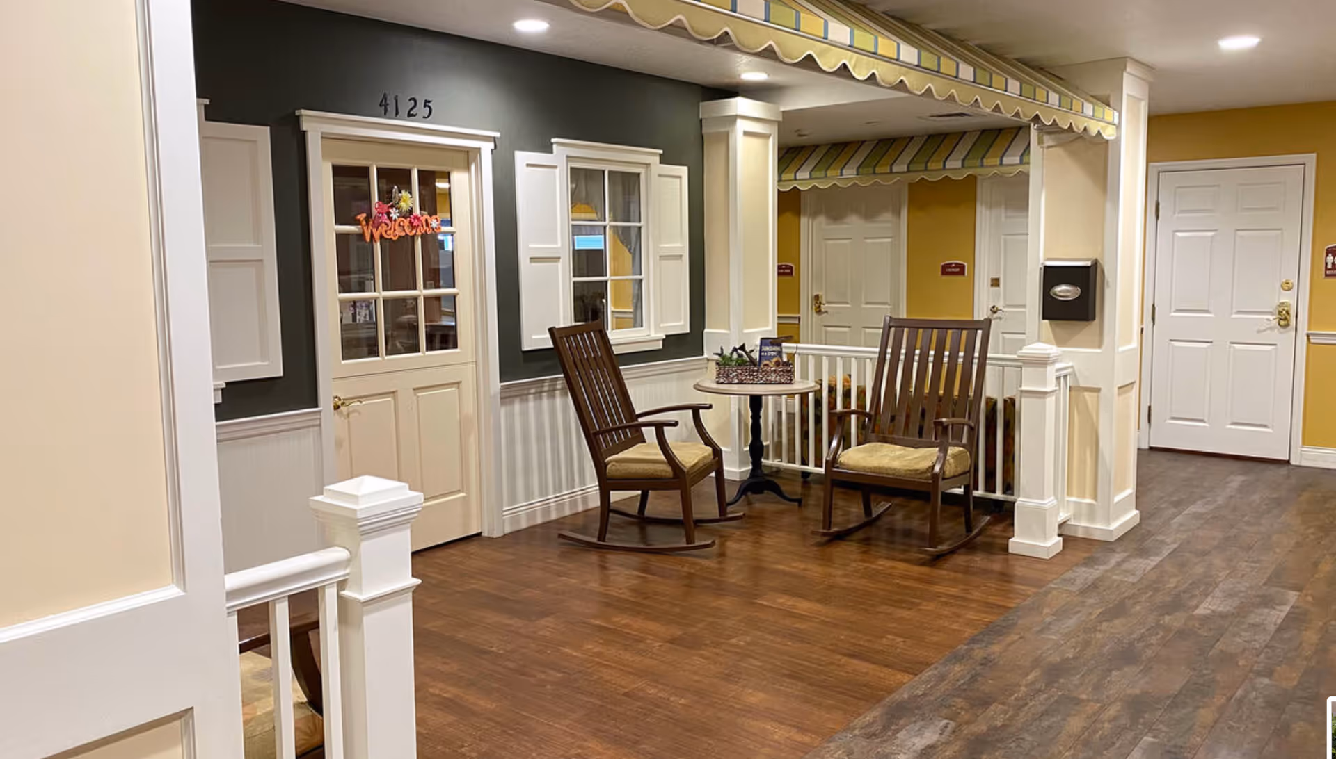 Interior corridor seating area with two wooden rocking chairs and a small table in front of a mock house facade bearing a 'Welcome' sign.