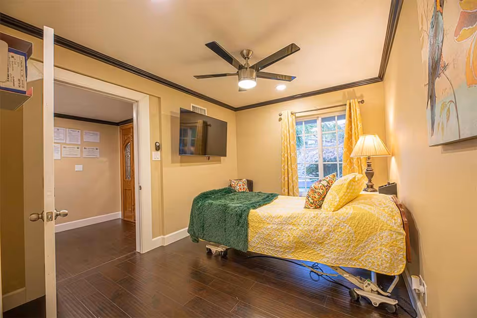 A cozy bedroom with a single bed covered in yellow and green bedding, a bedside lamp, a wall-mounted flat screen TV, a ceiling fan with lights, and a window with yellow curtains. The room has beige walls and dark wood flooring. An open door leads to another room with certificates on the wall.
