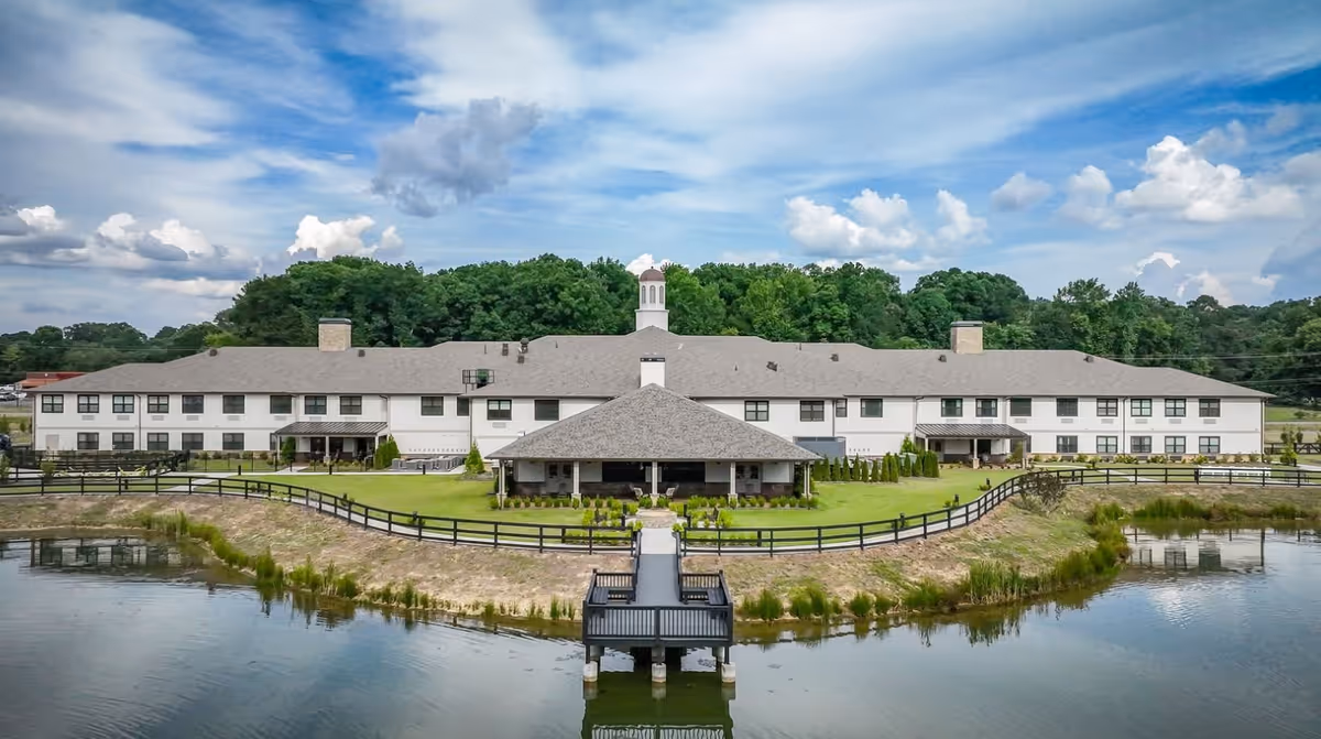 Wide exterior view of The Fountains of Calhoun building with a large pond in the foreground, a wooden dock extending into the water, green lawn, and a covered patio area. The building is two stories with a gray roof and white walls, surrounded by trees under a partly cloudy sky.