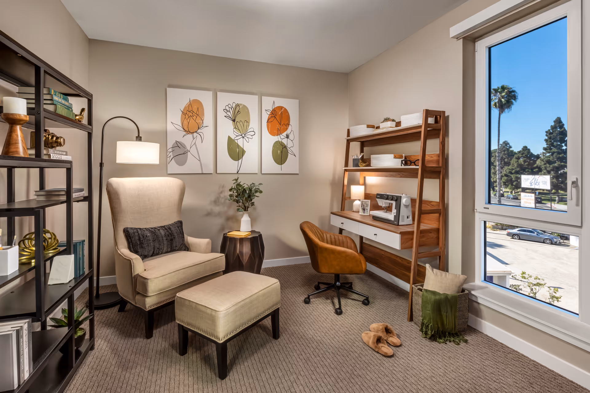 A cozy interior room featuring a beige armchair with a matching ottoman, a floor lamp, and a small side table with a vase of flowers. There is a wooden desk with a brown swivel chair, a sewing machine, and shelves with decorative items. A large window shows a view of trees and parked cars outside. The walls are decorated with three floral art prints.