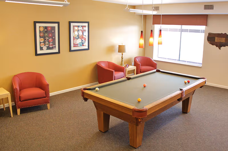 A well-lit room with a pool table in the center, surrounded by three red armchairs and a small wooden side table. The walls are beige with two framed pictures and a decorative wooden wall hanging. There is a window with blinds and three hanging pendant lights above the pool table.