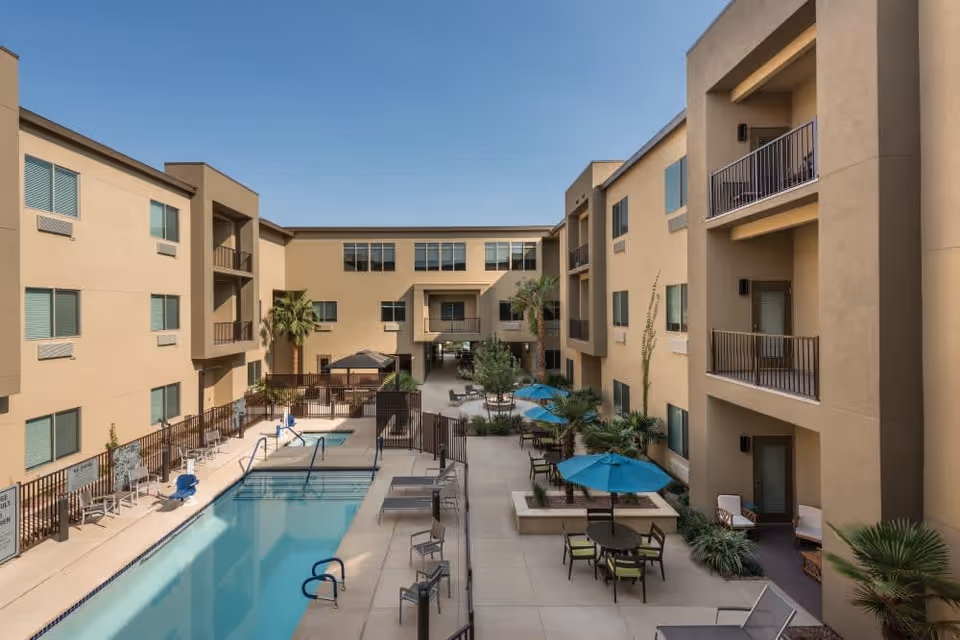 Open-air central courtyard of a multi-story senior living building featuring a swimming pool, patio seating with umbrellas, and balconies overlooking the space.