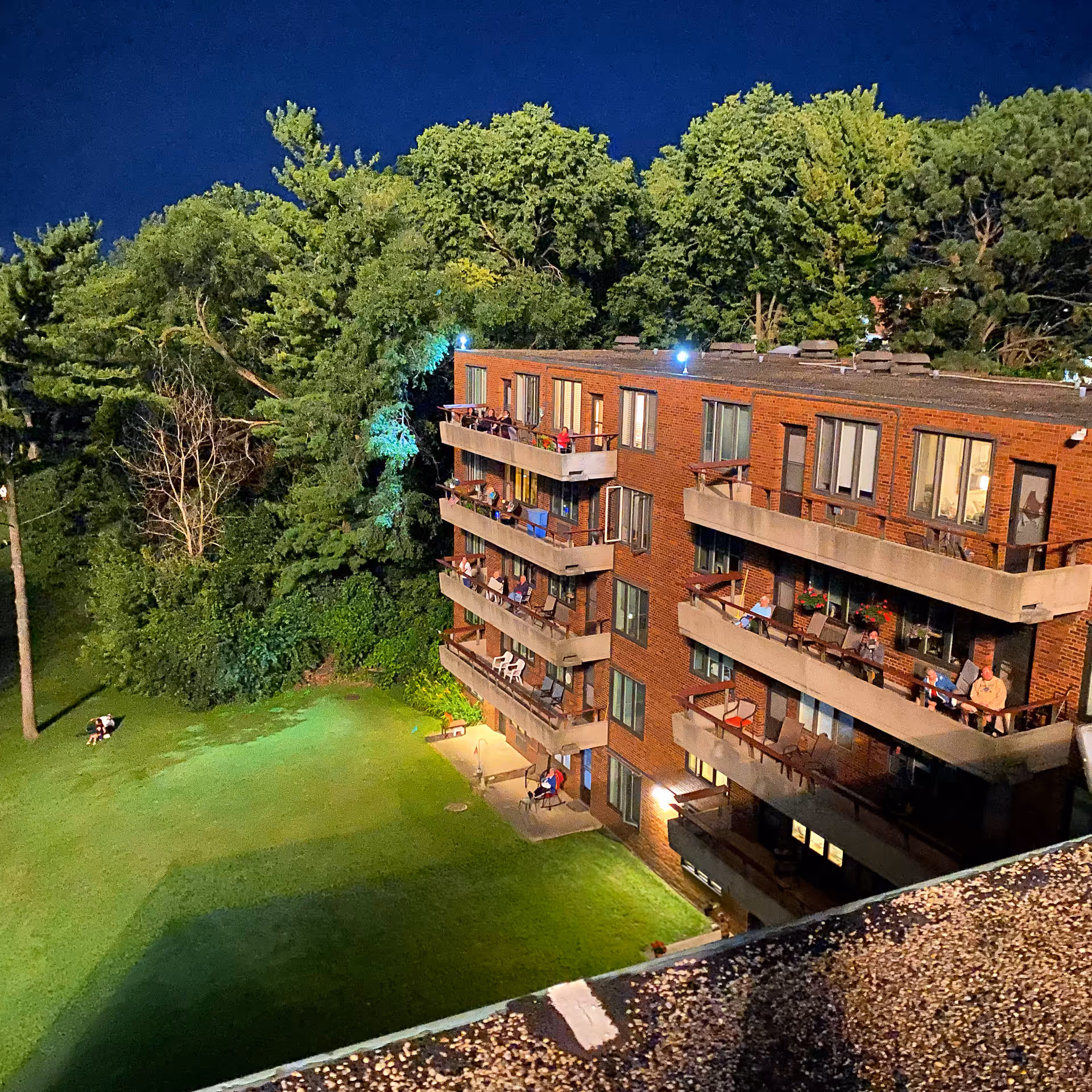 Nighttime view of a multi-story brick retirement community building with balconies where several people are sitting. The building is surrounded by a well-lit grassy area and dense trees in the background.
