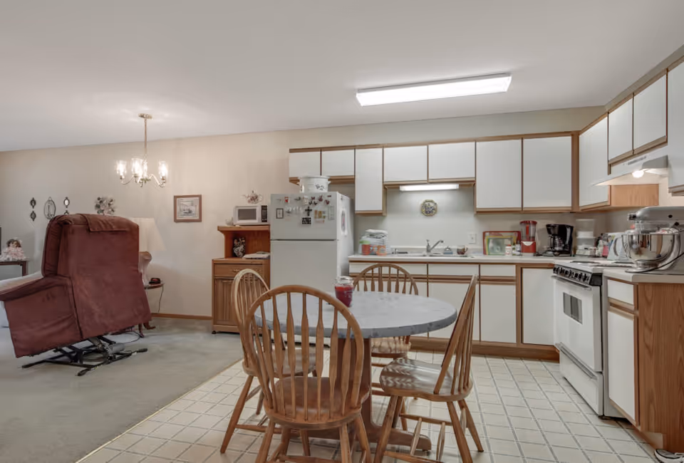 Interior view of a senior living facility kitchen and dining area at Keller Lake Commons. The kitchen features white cabinets with wooden trim, a white refrigerator, stove, and various kitchen appliances on the counters. In the foreground, there is a round dining table with four wooden chairs. To the left, a maroon recliner chair is positioned on a carpeted area adjacent to the kitchen. A chandelier hangs from the ceiling above the dining area.