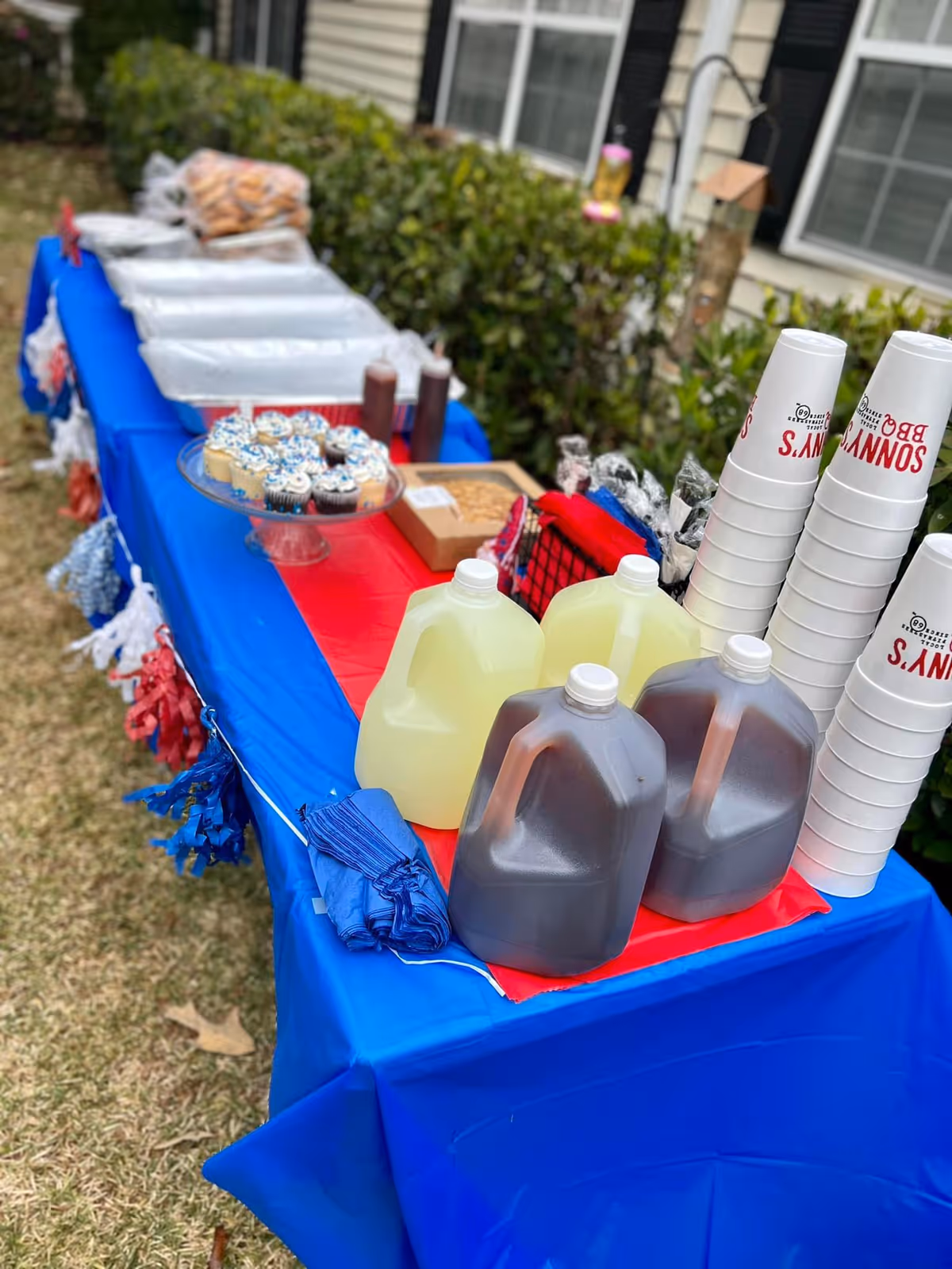An outdoor buffet table covered with red and blue tablecloths holding gallon jugs of drinks, stacked disposable cups, cupcakes and other food in front of a senior living building.