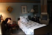 A cozy nursing home bedroom with a single bed covered in floral bedding, a wooden chair with cushions, a small side table with a lamp and decorative items, and a comfortable armchair. The walls are decorated with framed pictures and a wreath.